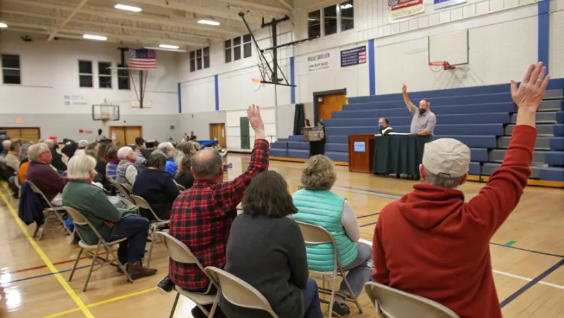 Residents attending a New Hampshire town meeting in an elementary school gymnasium, raising hands to vote on a local issue